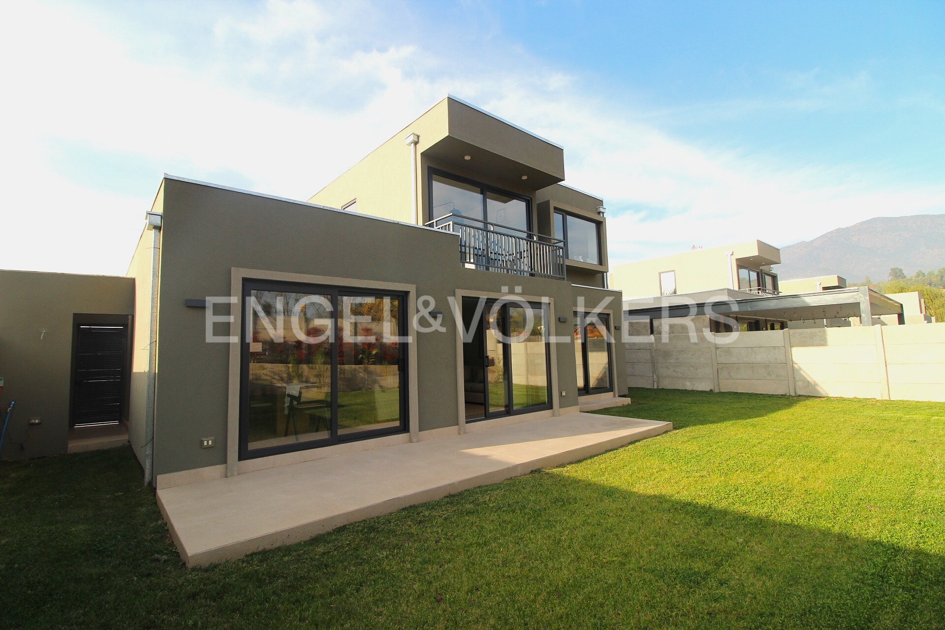 Two-story modern house with a balcony, large windows, and a green lawn under a blue sky. Engel & Völkers logo on the windows.