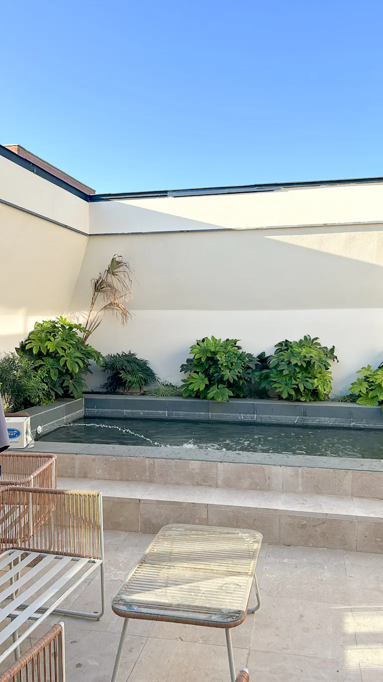 Rooftop patio with a shallow pool, green plants, and white walls under a clear blue sky. A table and chairs sit on the stone patio.