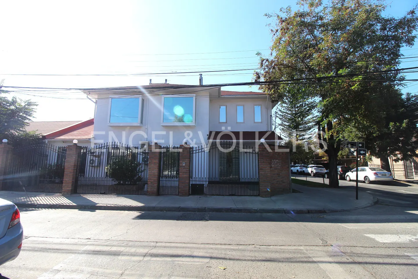 Two-story white house with a black iron fence and brick pillars, Engel & Völkers logo, and street view.