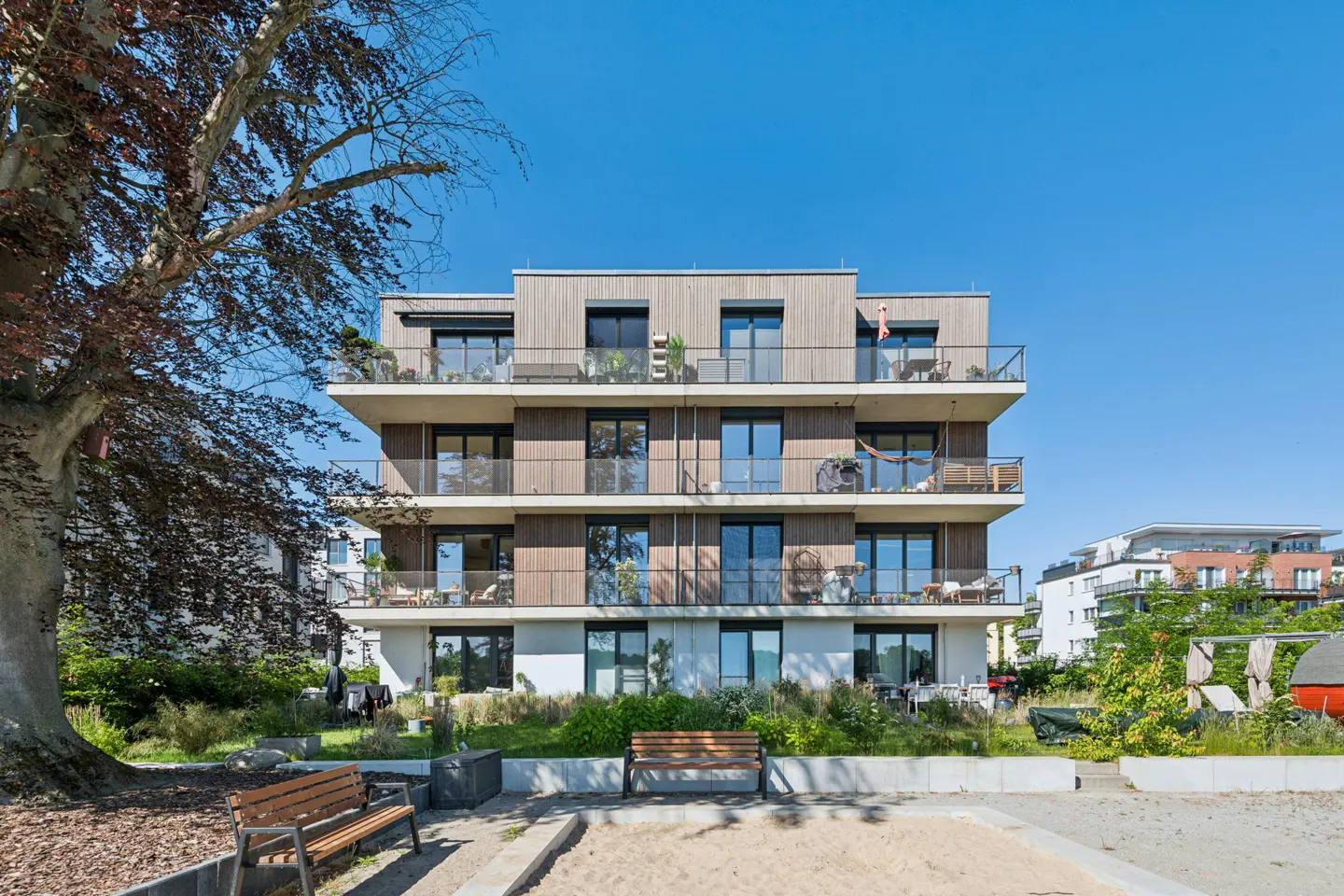 Modern four-story apartment building with wood-paneled balconies, a sandy playground, and benches under a clear blue sky.