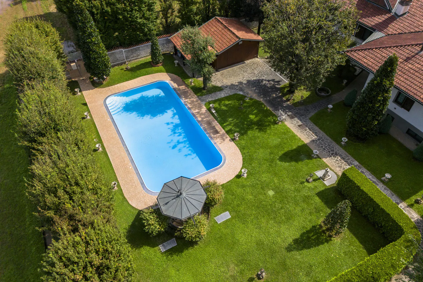 Aerial view of a rectangular pool with blue water, surrounded by green grass, trees, and a gazebo in a backyard.