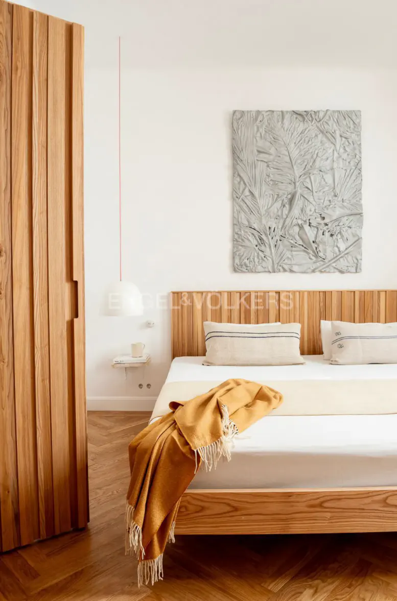 Bedroom with wooden bed frame, headboard, and wardrobe. A yellow blanket is draped on the bed. A gray art piece hangs above the bed.