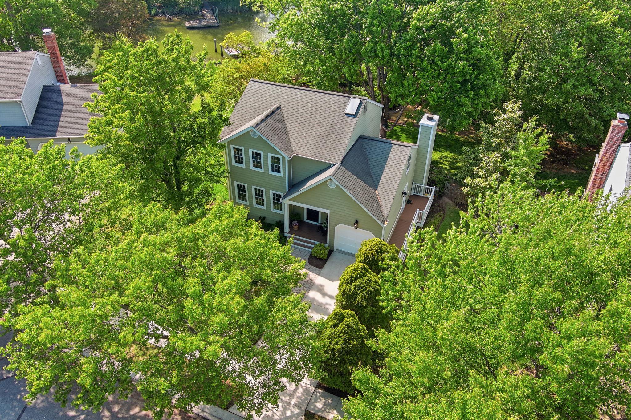 Aerial view of a two-story, light green house with a gray roof, surrounded by lush green trees and a glimpse of a river in the background.