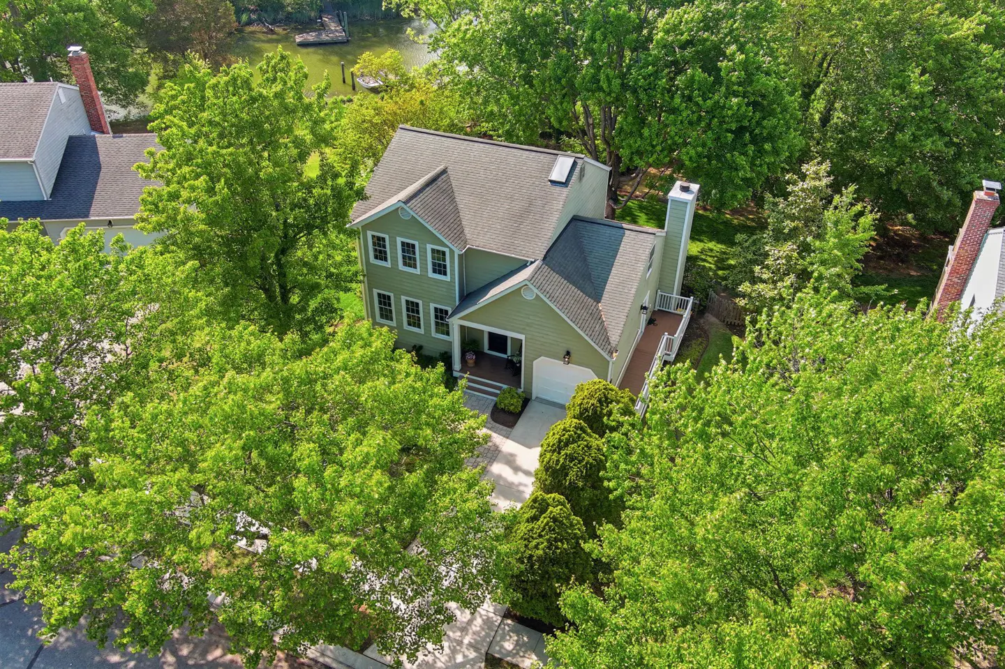Aerial view of a two-story, light green house with a gray roof, surrounded by lush green trees and a glimpse of a river in the background.