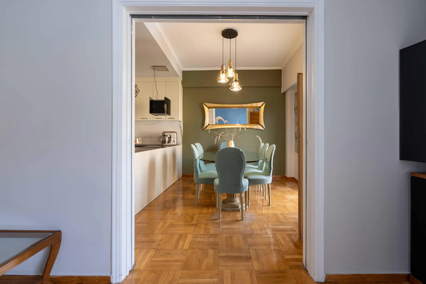 View through doorway into dining room with parquet floors, green walls, and a table with six light blue chairs. A kitchen is visible in the background.