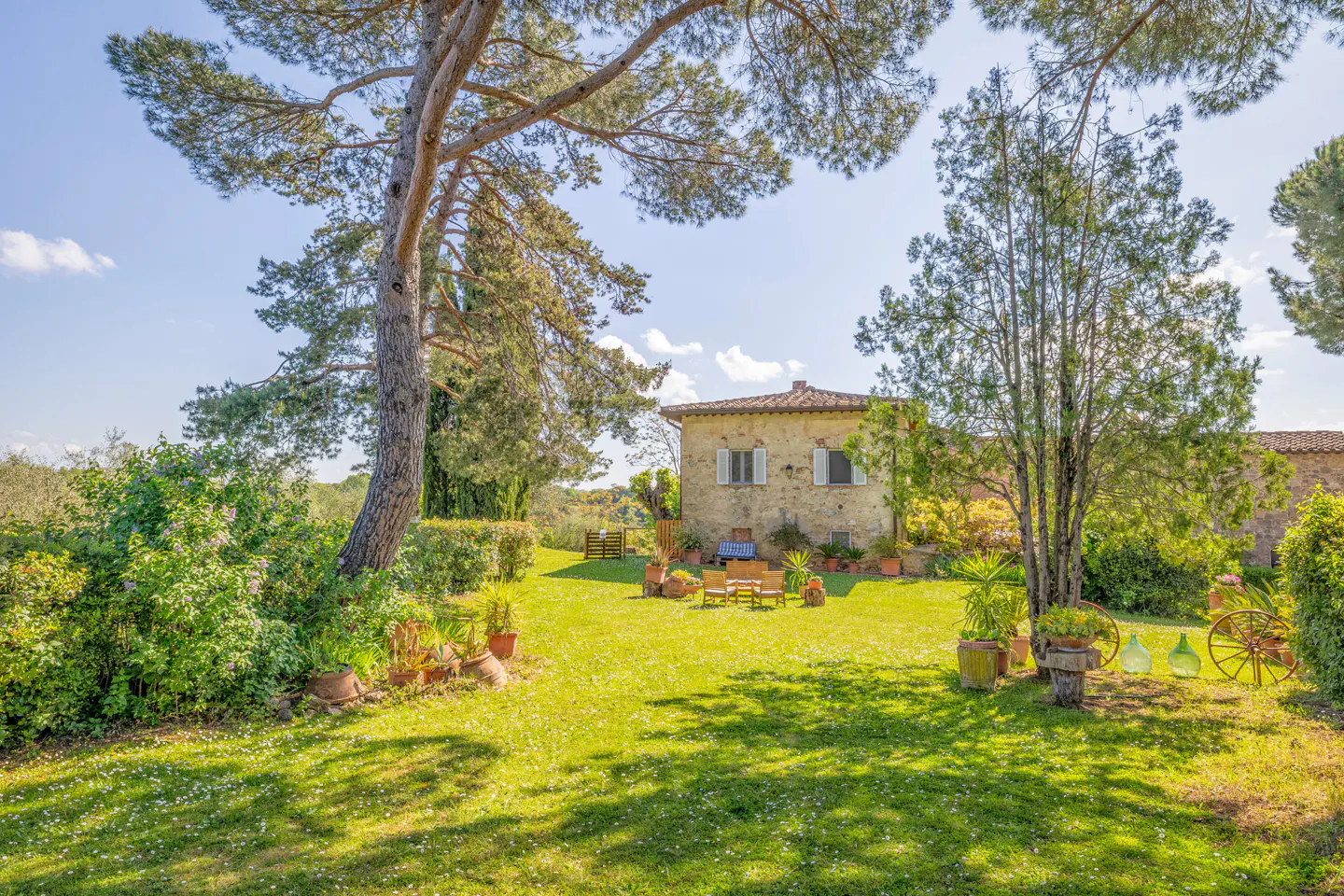 Exterior view of a stone house with white shutters, surrounded by a green lawn, trees, and potted plants on a sunny day.
