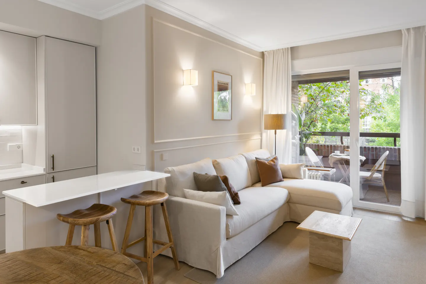 Living room with white sofa, wood stools at a white counter, and balcony with table and chairs.