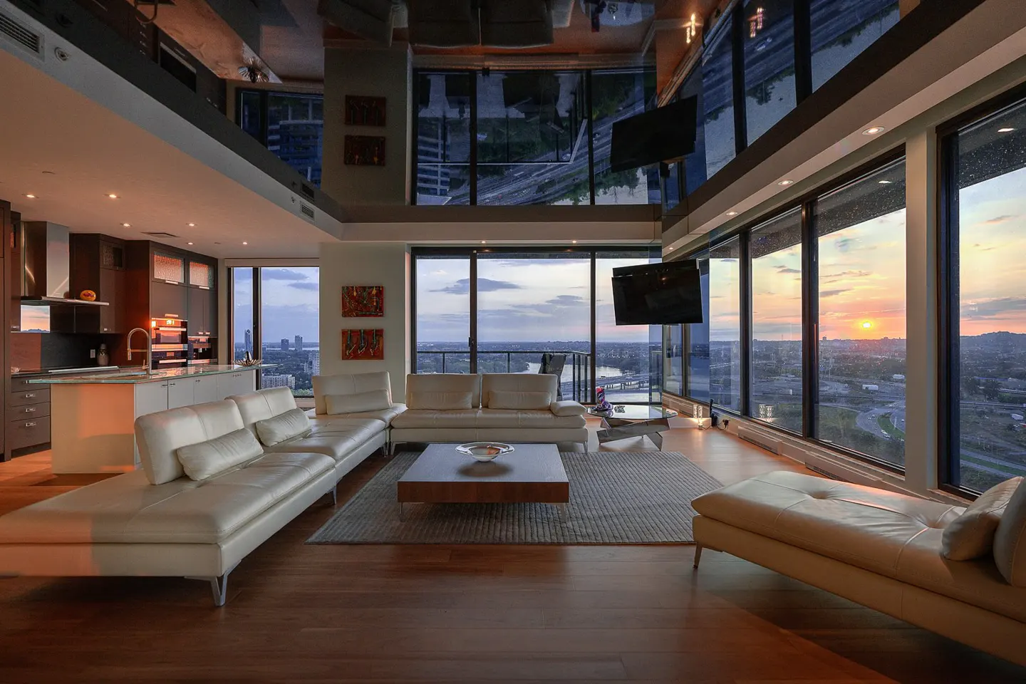 A modern condo interior with white sofas, wood floors, and floor-to-ceiling windows showing a sunset city view.