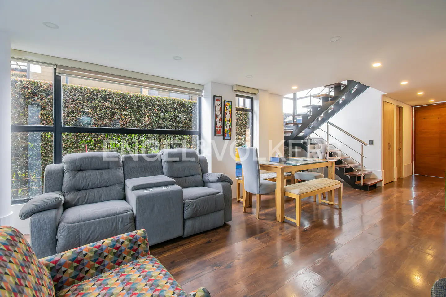 Living room with gray reclining sofa, wood table and bench, and black metal stairs. Large window with green foliage outside.