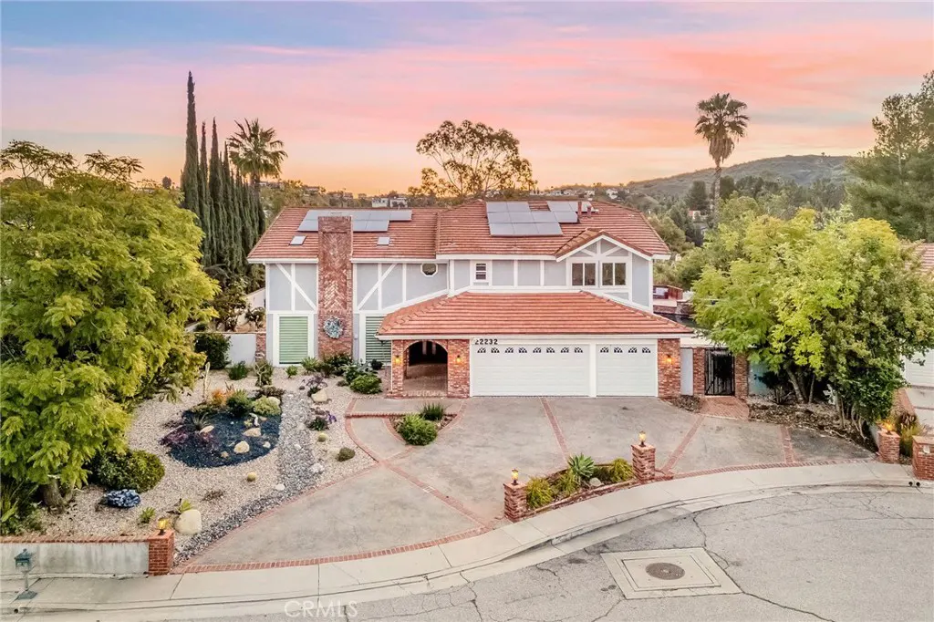 Two-story house with a red tile roof, white garage doors, and a gray exterior at sunset. Solar panels are visible on the roof.