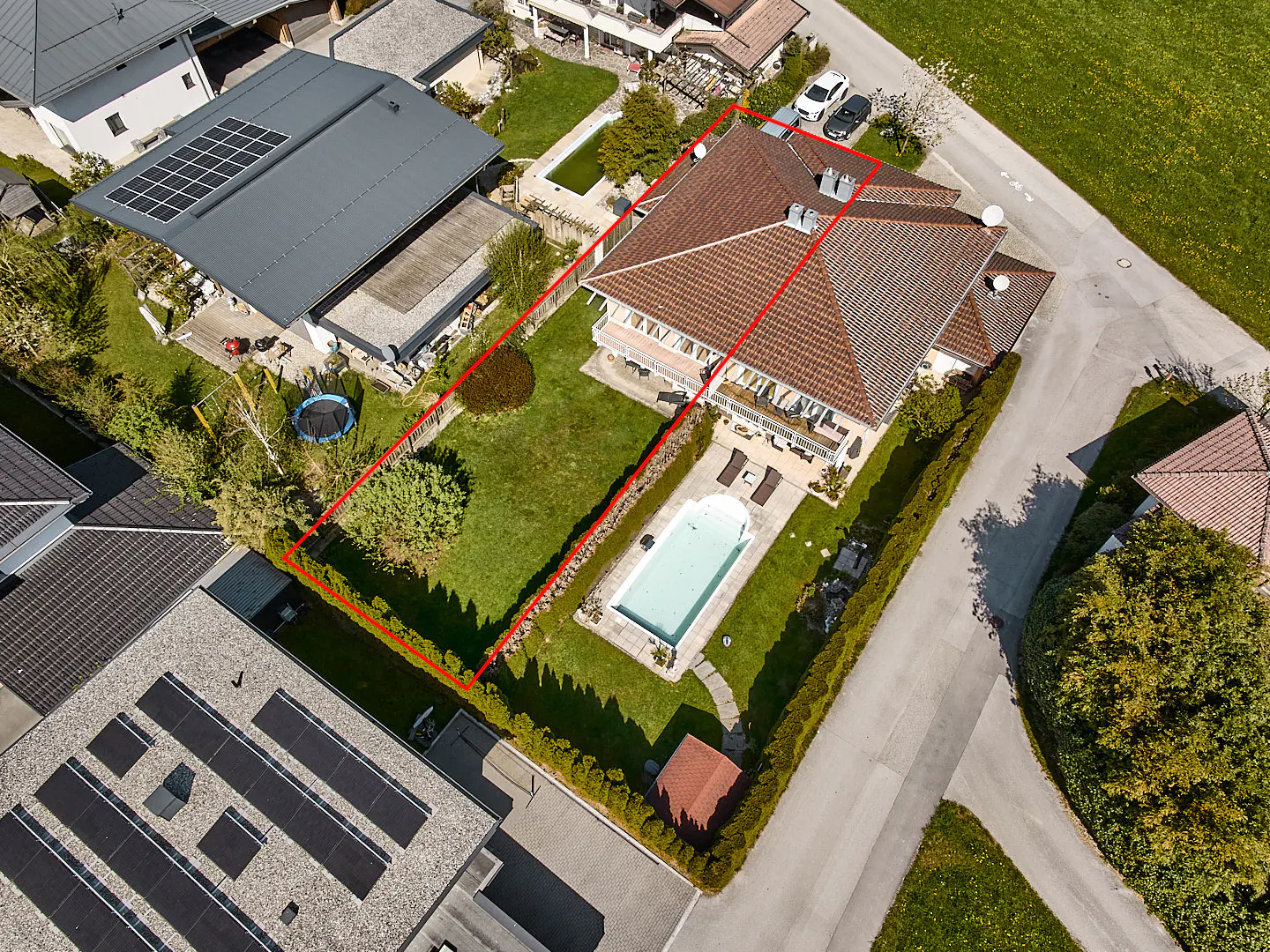 Aerial view of a house with a red tile roof, a pool, and a green lawn, surrounded by a red boundary line.