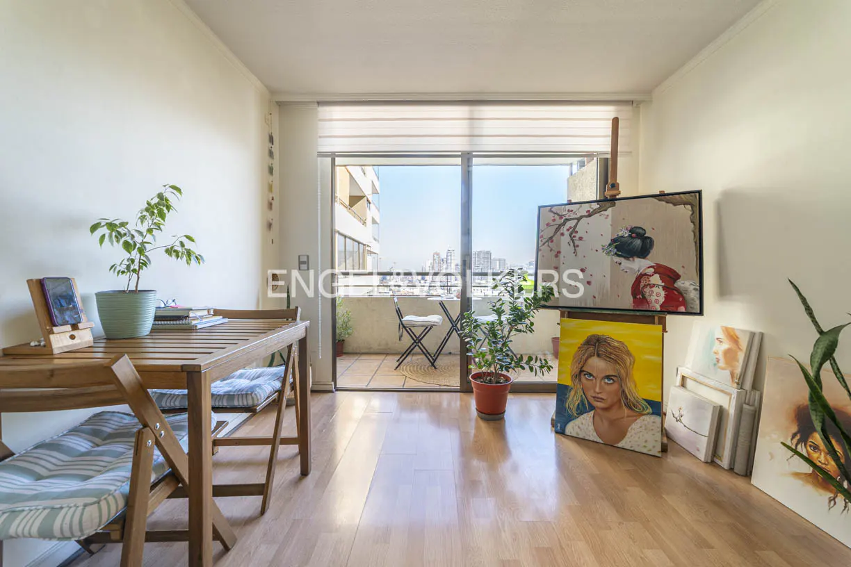 Bright apartment interior with wooden floors, table, chairs, and balcony. Paintings lean against the wall, adding an artistic touch.