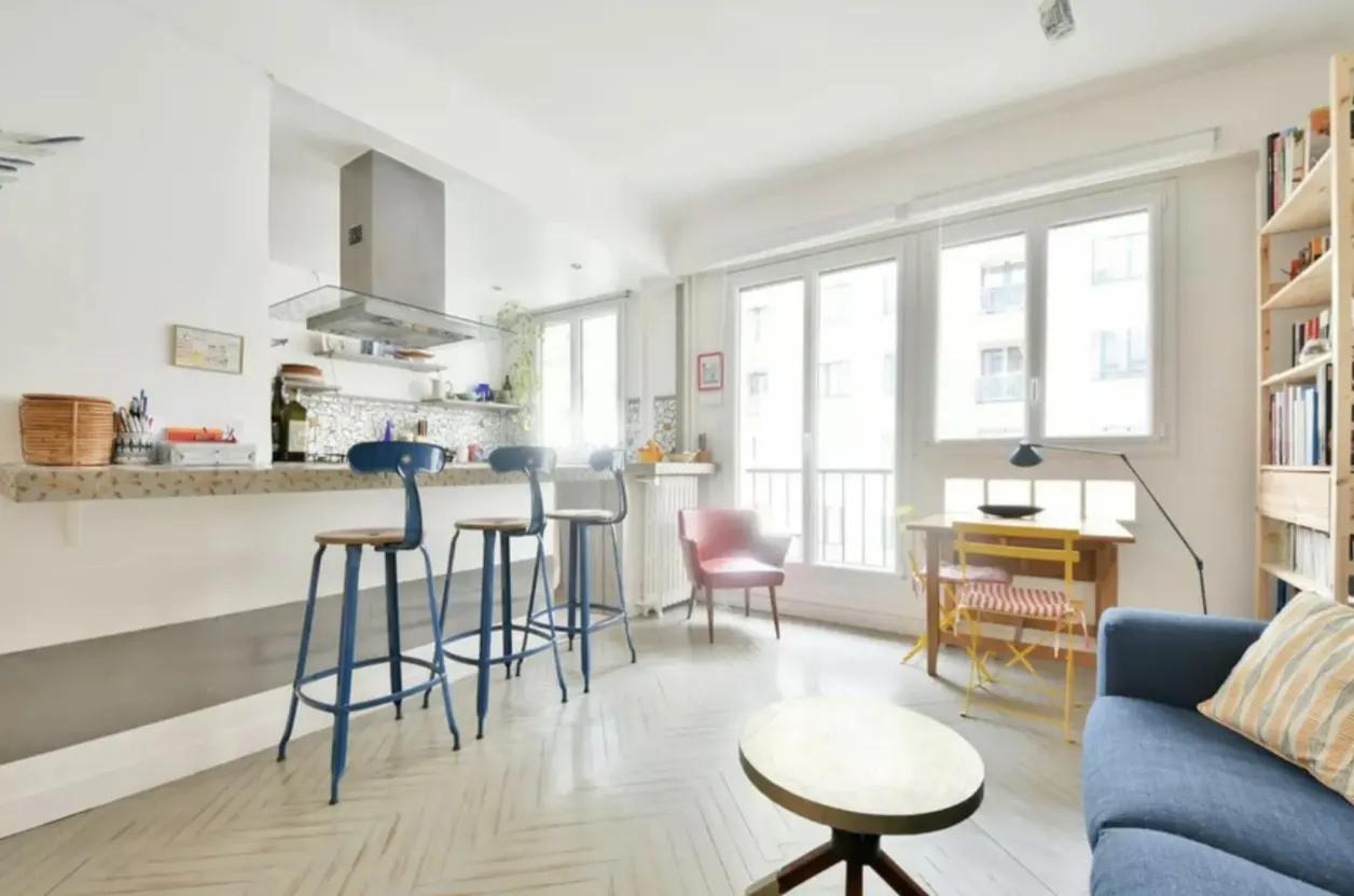 Bright, open apartment with a kitchen bar, blue stools, and a herringbone floor. A blue sofa and colorful chairs add pops of color.