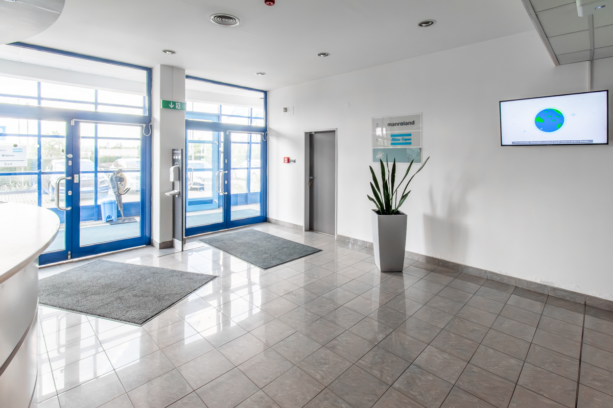 Bright office lobby with tiled floors, glass doors, a potted plant, and a wall-mounted screen displaying a globe graphic.