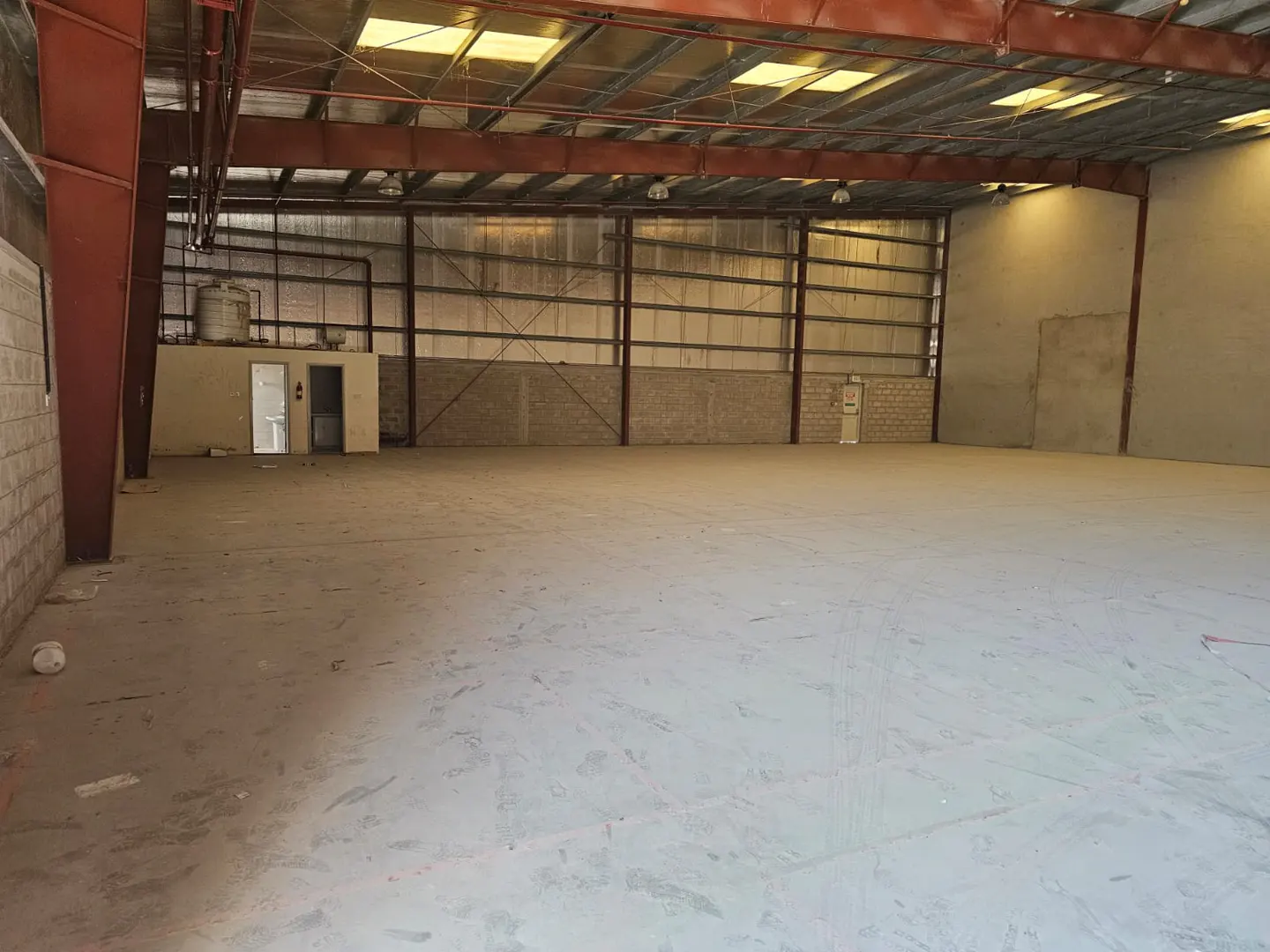 Empty warehouse interior with concrete floor, metal beams, and skylights. Doors and a water tank are visible in the background.