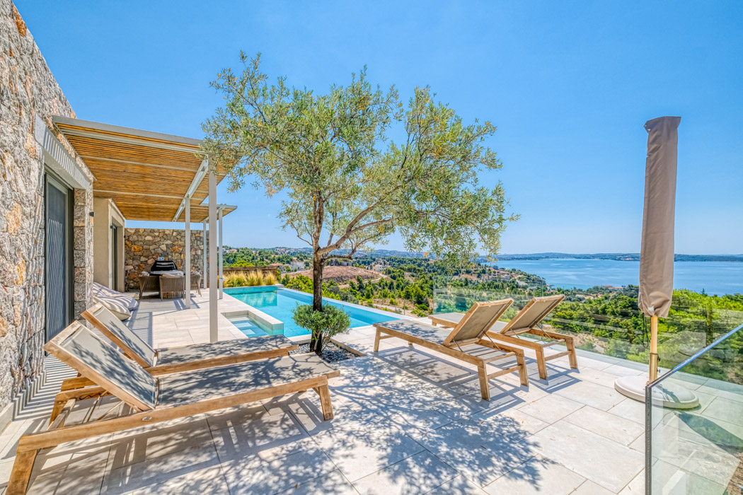 Stone patio with wooden lounge chairs, an olive tree, and an infinity pool overlooking the sea under a clear blue sky. [AI-generated text]