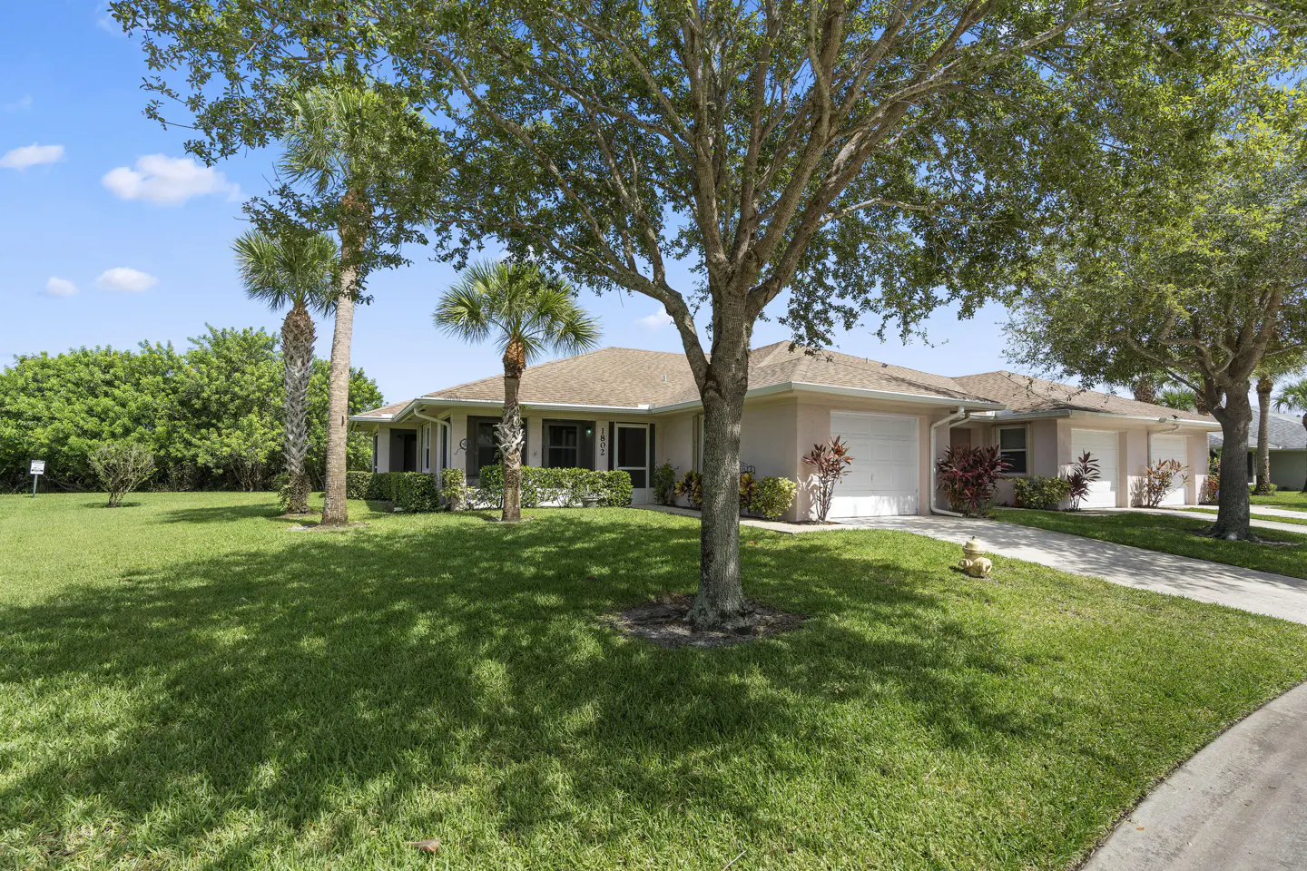 Single-story beige house with a tan roof, green lawn, palm trees, and a concrete driveway under a clear blue sky.