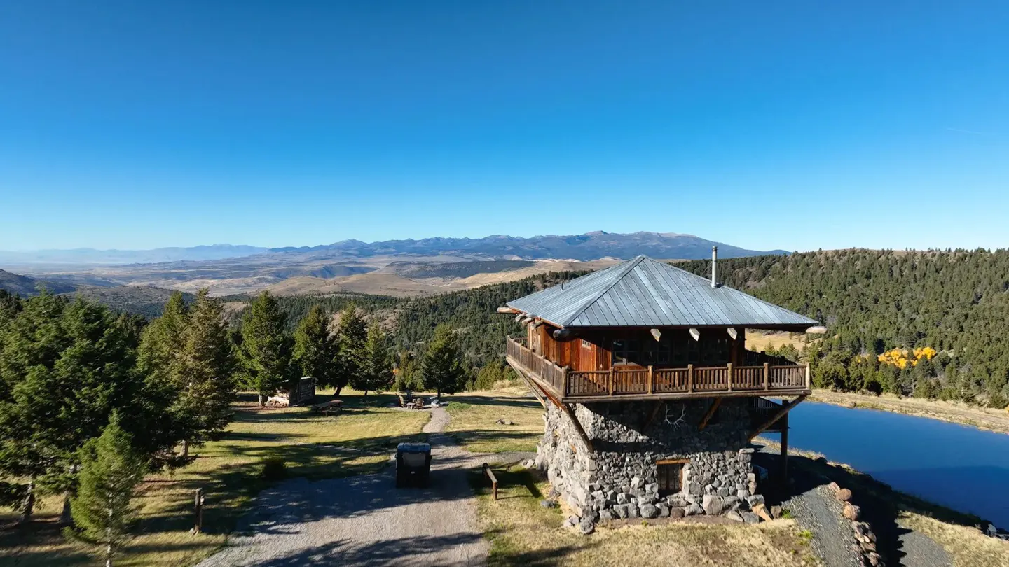 Two-story stone tower with a metal roof and wooden balcony overlooking a lake and mountain range under a clear blue sky.