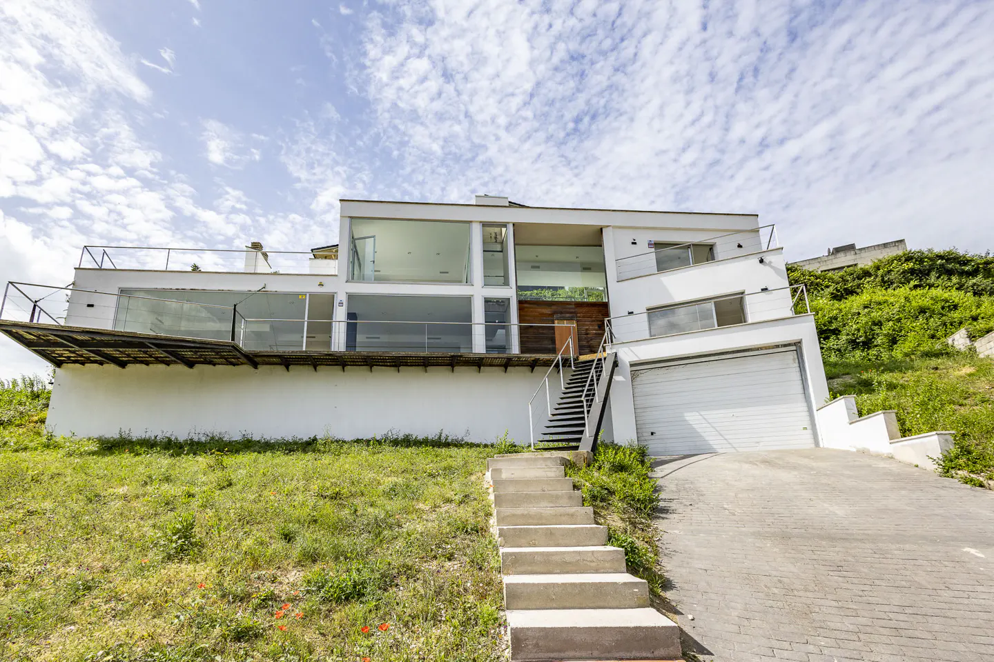 Modern white house on a grassy hill with a garage, stairs, and large windows under a cloudy sky.