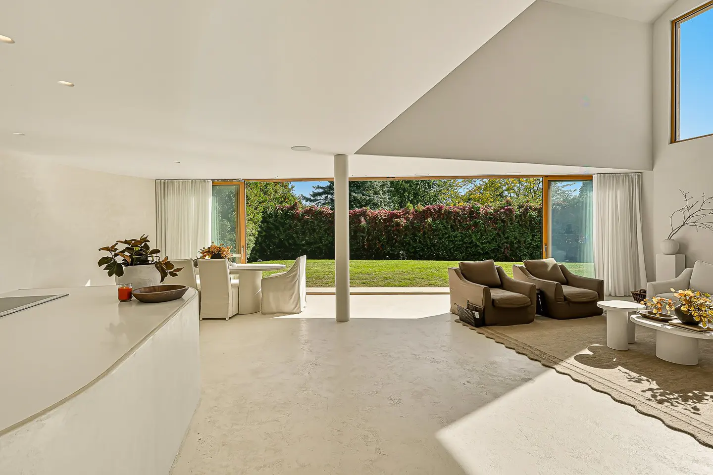 Bright, open-concept living space with white walls, a dining table, and two brown armchairs. Large windows offer a view of the green lawn and trees.