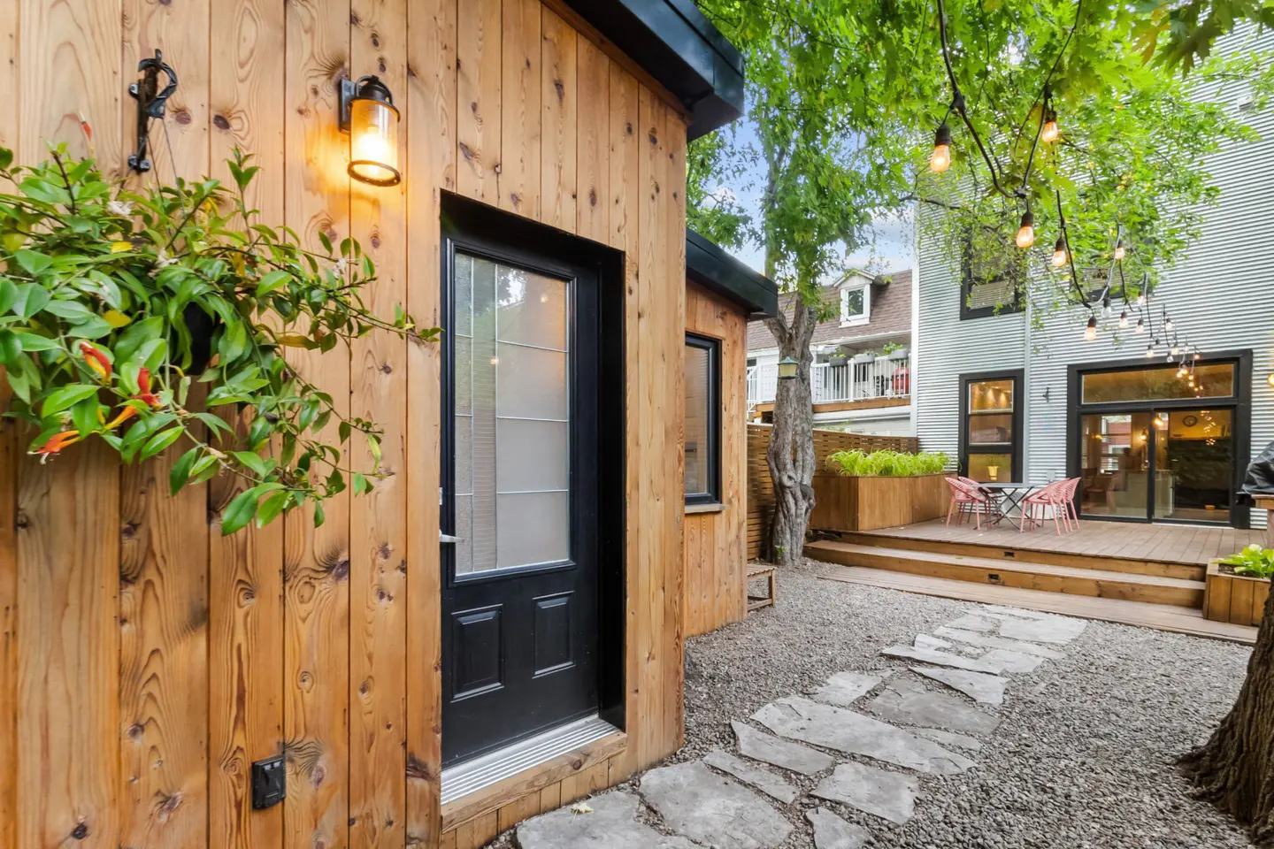 Exterior view of a wood-paneled house with a black door, a stone path, and a backyard patio with string lights.