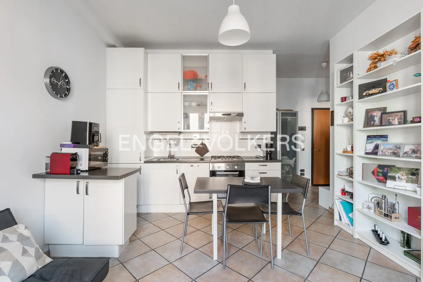 A bright kitchen with white cabinets, a black table with four chairs, and a bookshelf filled with decorative items.