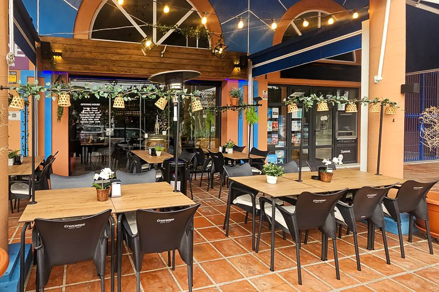 Outdoor restaurant patio with wooden tables, black chairs, and terracotta tile flooring. String lights and plants decorate the space.
