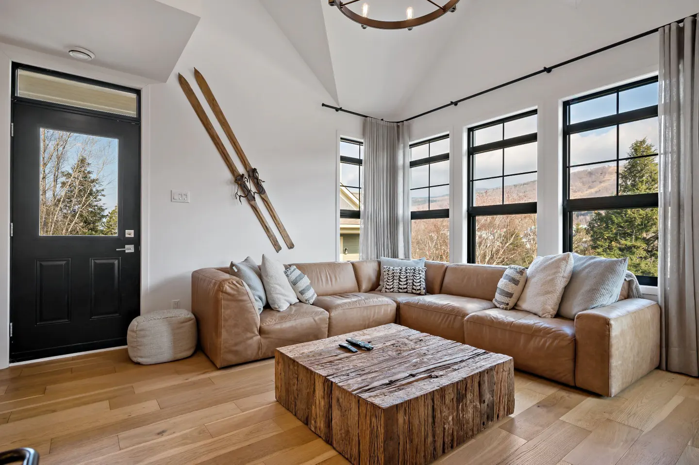 Living room with a brown leather sectional sofa, rustic wood coffee table, and vintage skis on the wall. Large windows offer a view of trees.
