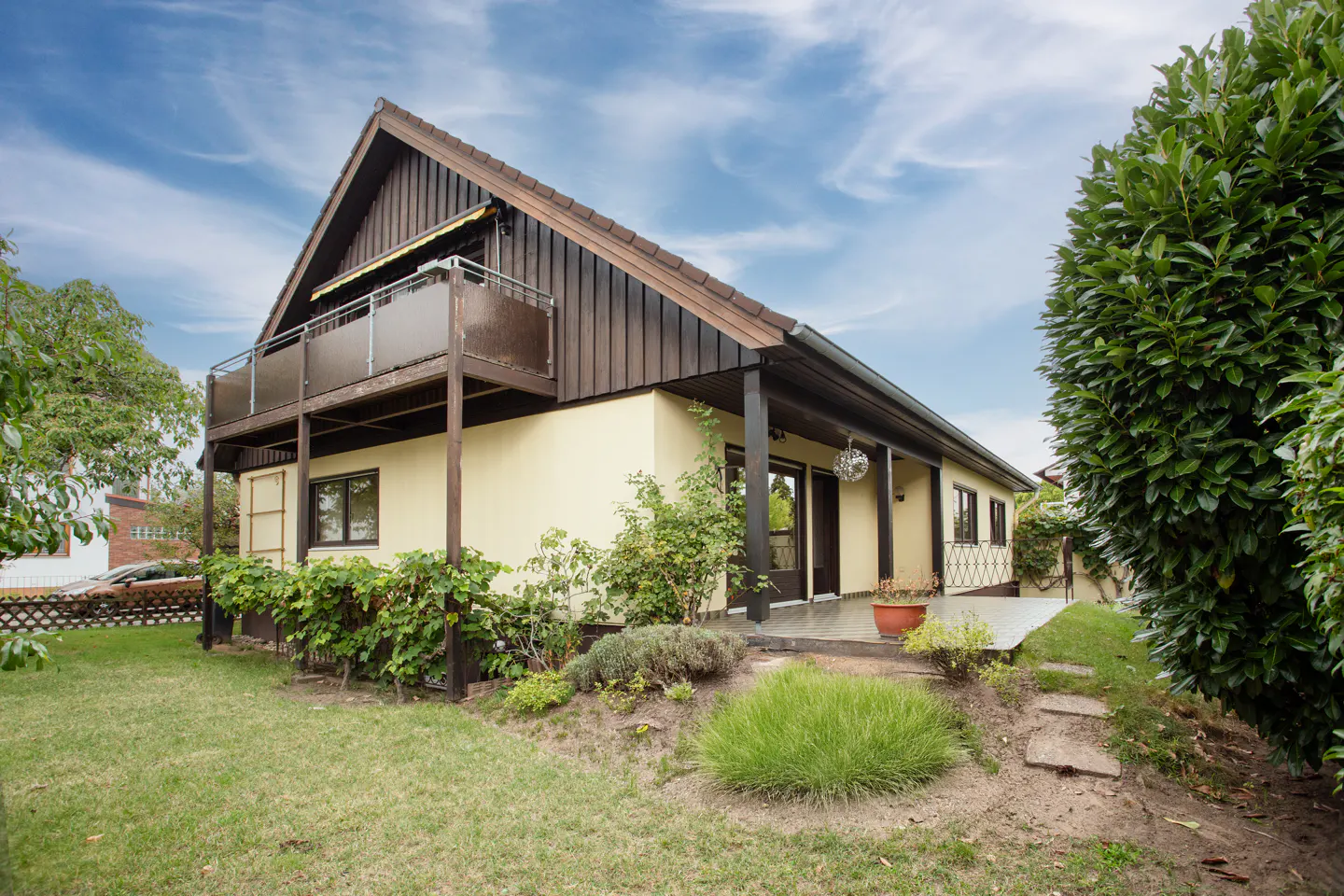 Two-story house with a balcony, dark brown trim, and a light yellow exterior, surrounded by green grass and foliage.