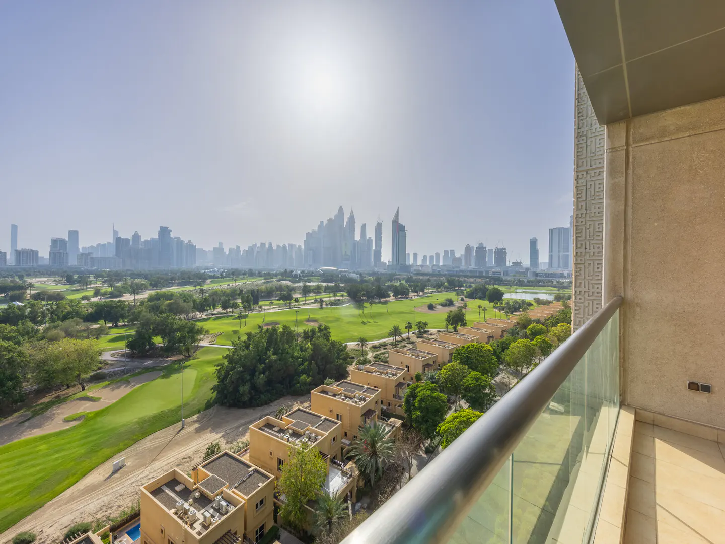 Balcony view of Dubai skyline, golf course, and tan villas. Metal railing in foreground. Sunny day.