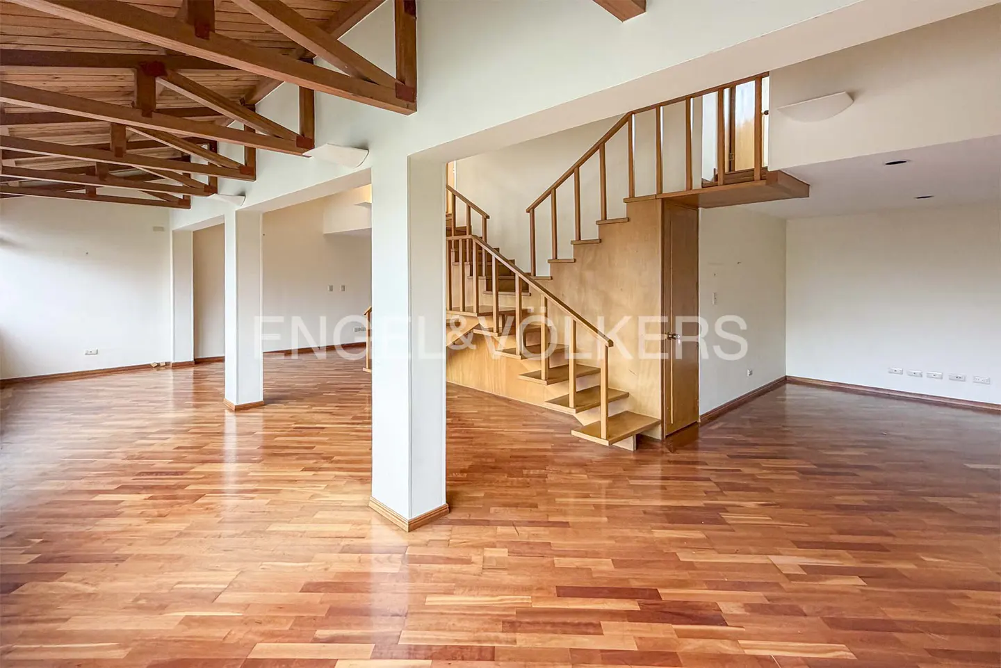 An interior view of a spacious room with hardwood floors, white walls, and wooden stairs. Exposed wooden beams are visible on the ceiling.
