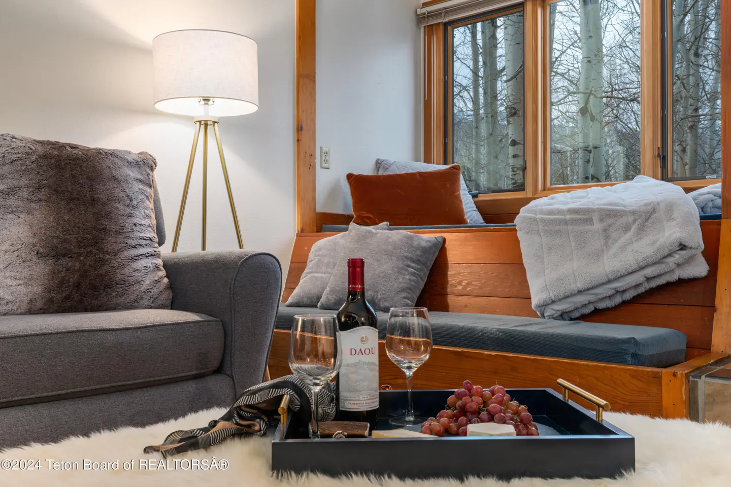 Cozy living room with a gray sofa, wine, grapes, and a window seat overlooking a snowy forest.