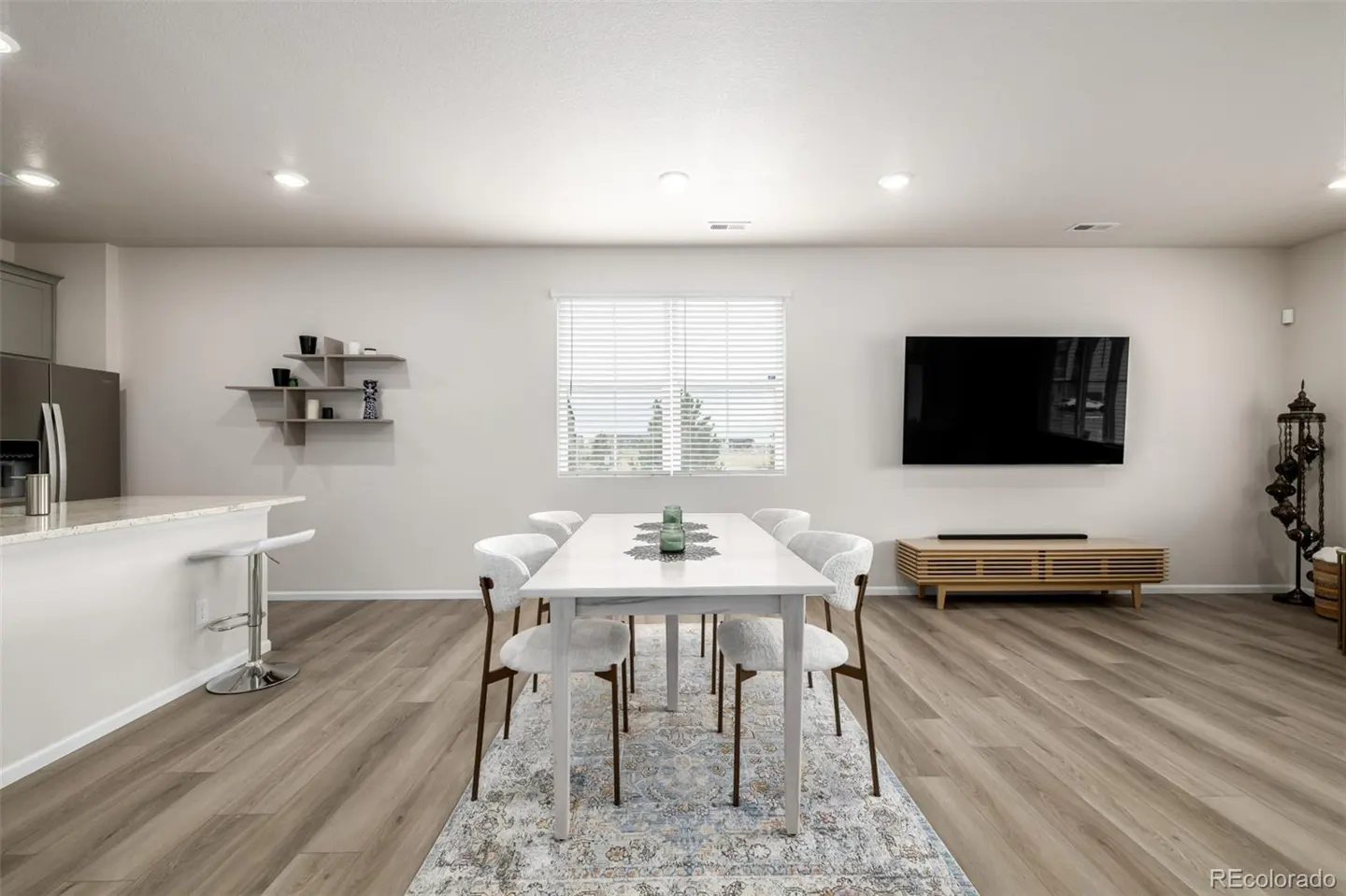 Open-concept dining area with a white table, six chairs, and a patterned rug on a wood floor. A TV is mounted on the wall.