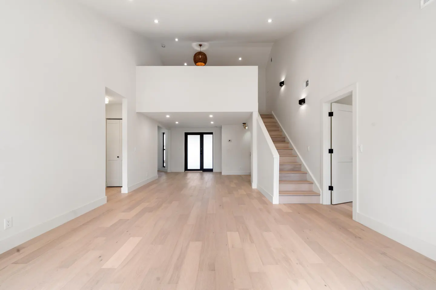 Bright, empty living room with white walls, light wood floors, and a staircase leading to a loft area with a woven pendant light.