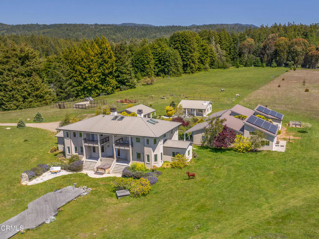 Aerial view of a large, gray house with a metal roof, surrounded by green lawns, trees, and outbuildings on a sunny day. A horse grazes nearby.