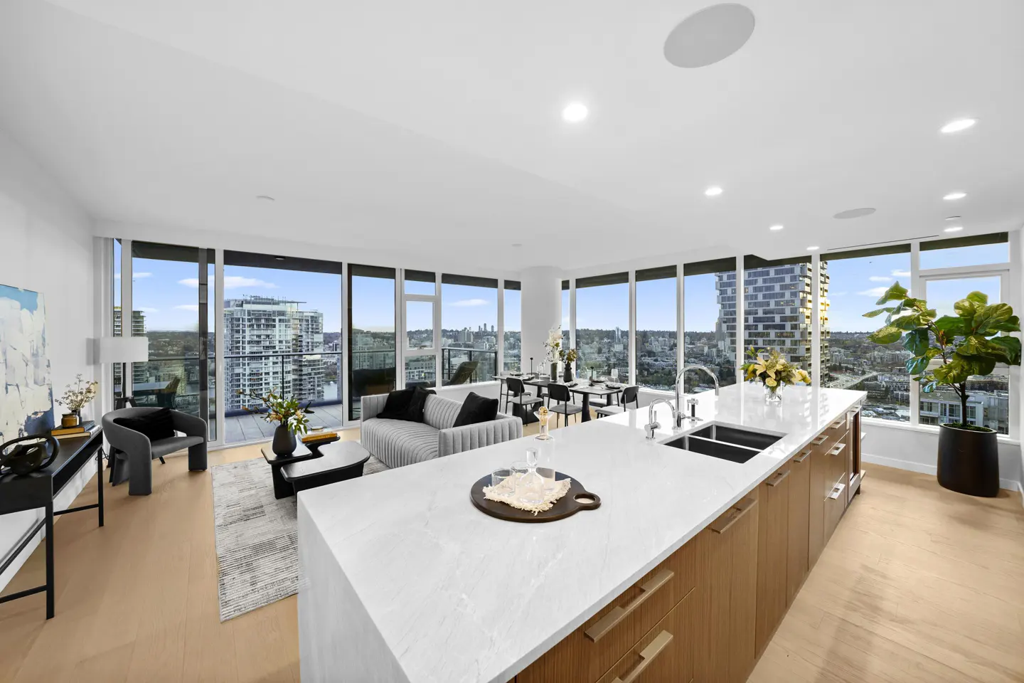 Bright, modern condo interior with floor-to-ceiling windows overlooking a city skyline. A kitchen island with white countertop and wood cabinets is in the foreground.