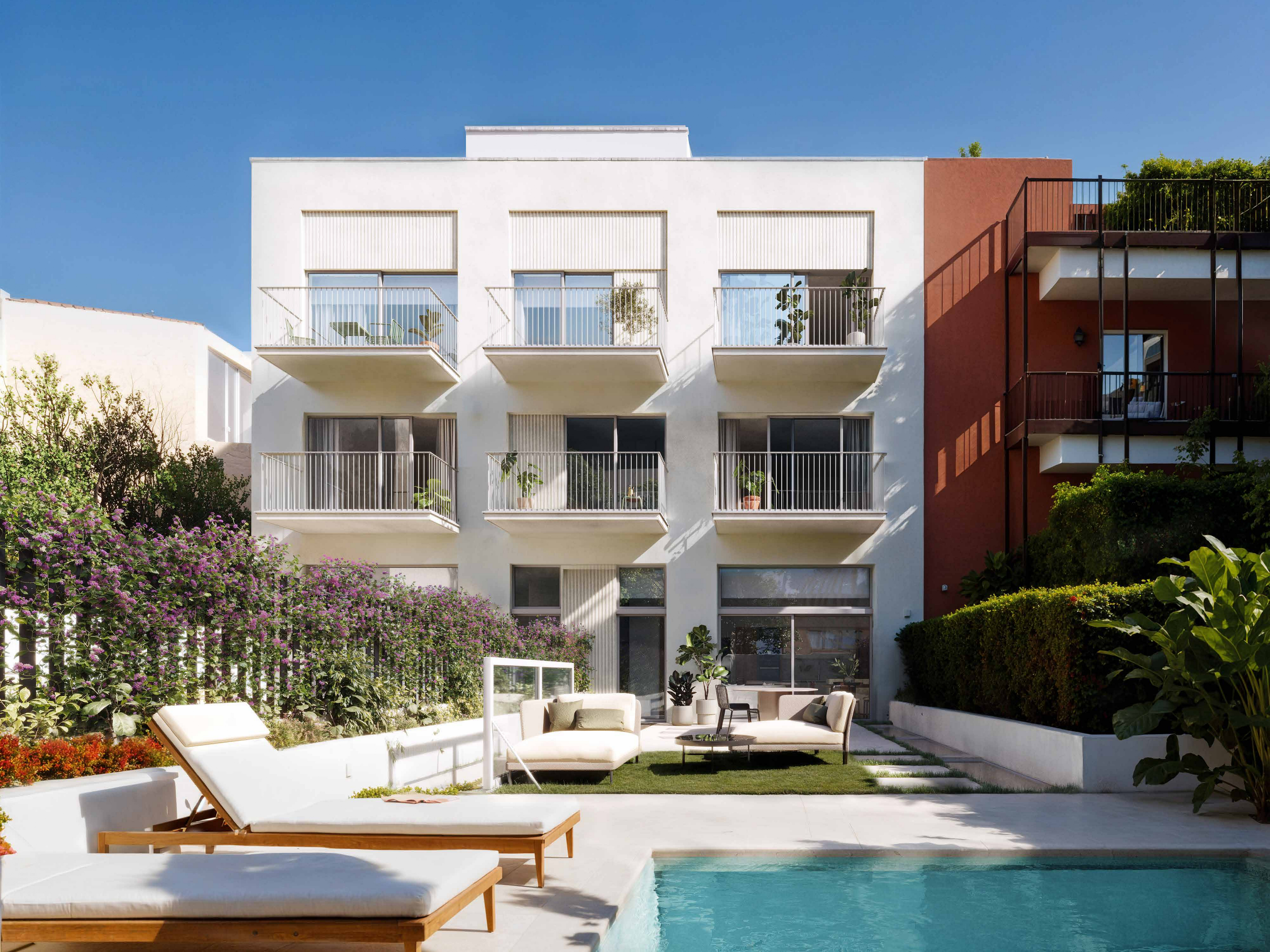 Exterior view of a modern white building with balconies, a pool, and lounge chairs under a clear blue sky.
