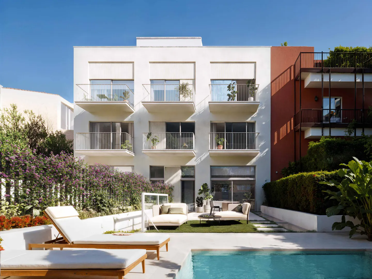 Exterior view of a modern white building with balconies, a pool, and lounge chairs under a clear blue sky.