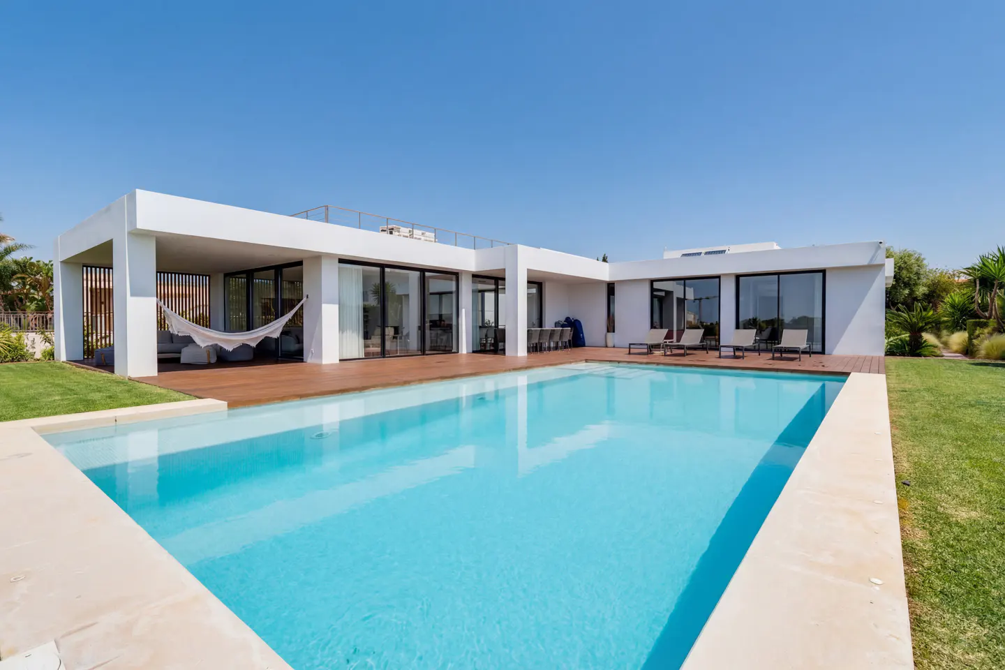 Modern white house with a blue swimming pool, wooden deck, and green lawn under a clear blue sky.