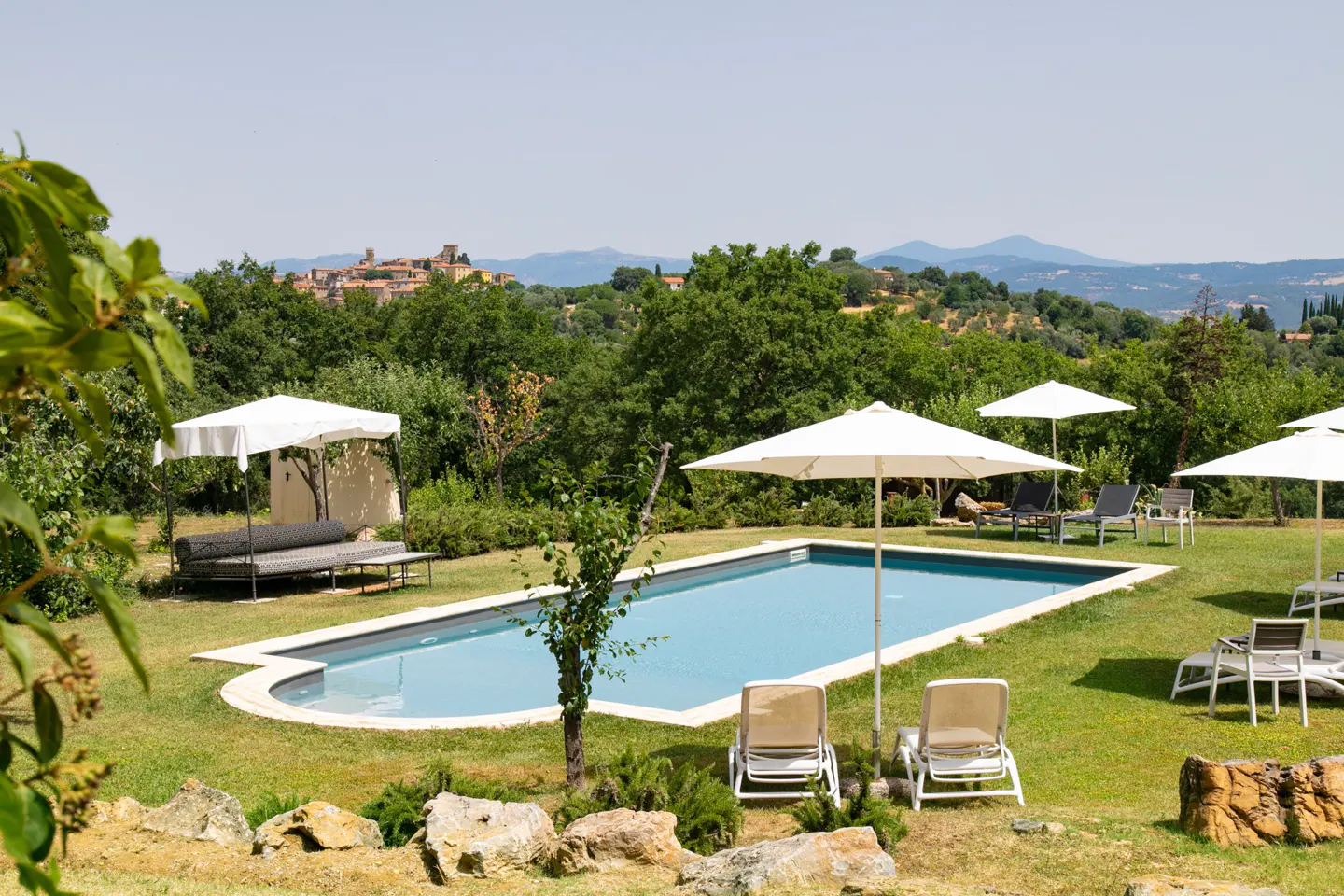 Rectangular pool with lounge chairs and umbrellas on green lawn. Trees and a distant town are in the background.
