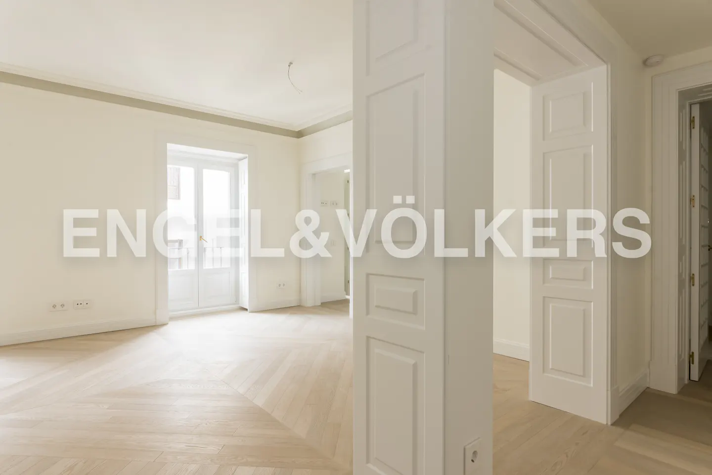 Bright, empty room with herringbone wood floors, white walls, and white trim. Engel & Völkers logo overlay.