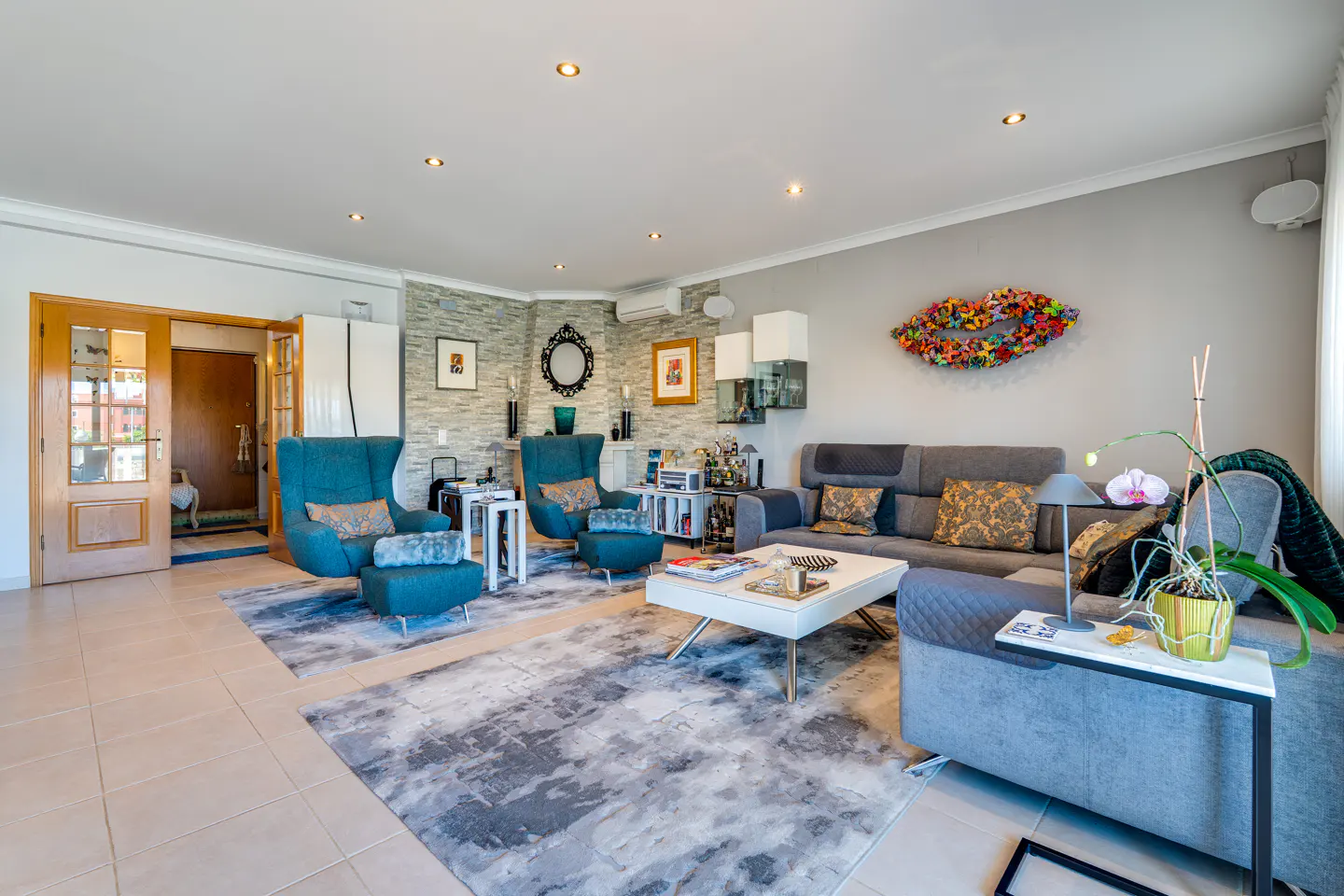Living room with gray sofas, teal chairs, and a gray and white rug. A white coffee table sits in the center. Artwork adorns the gray walls.