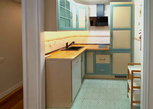 A compact kitchen with blue and cream cabinets, a wooden countertop, and patterned blue flooring. A black sink and appliances are visible.