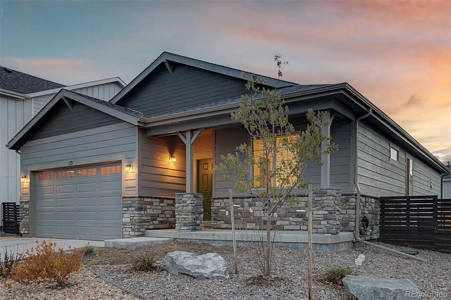 A gray, single-story house with stone accents and a covered porch at dusk. A young tree stands in the gravel yard.