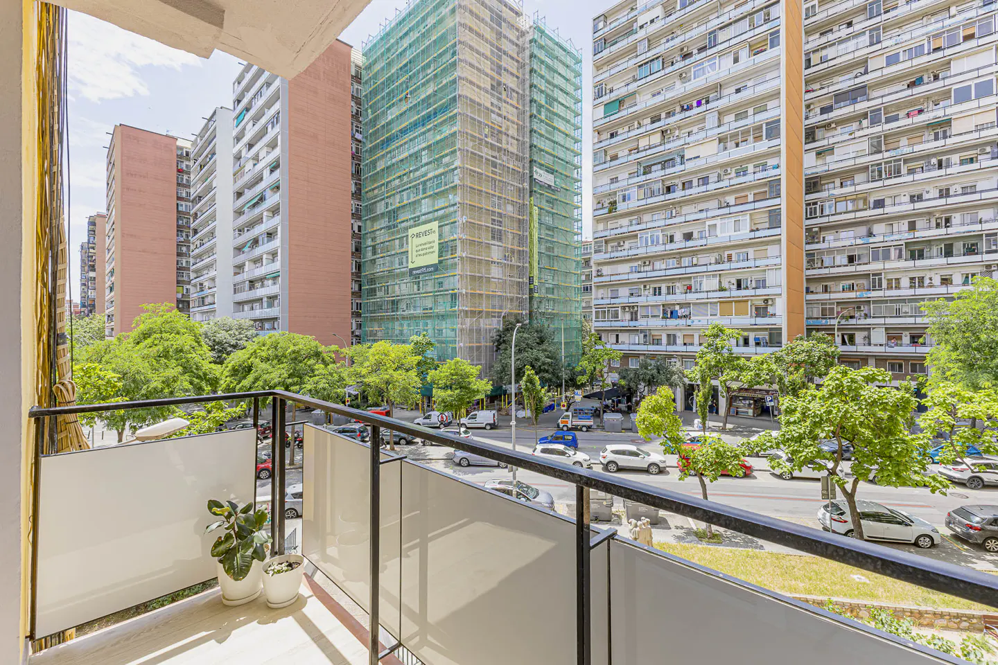 View from a balcony overlooking a city street with tall apartment buildings, green trees, and parked cars.