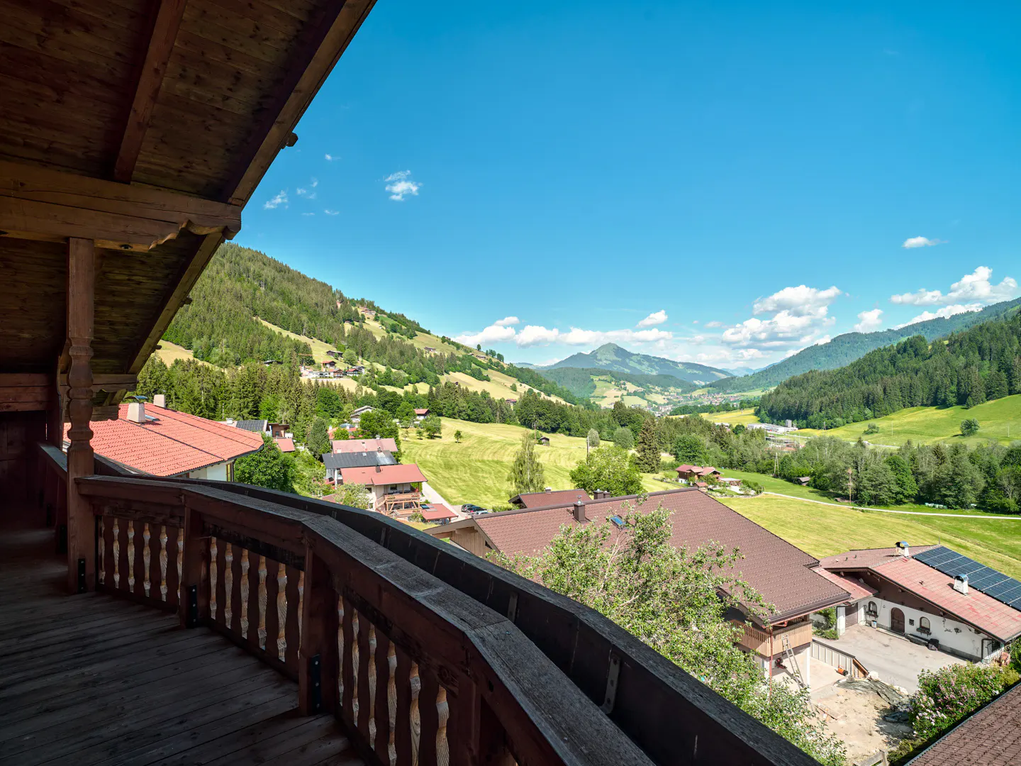 View from a wooden balcony overlooking a lush green valley with houses, rolling hills, and a clear blue sky.