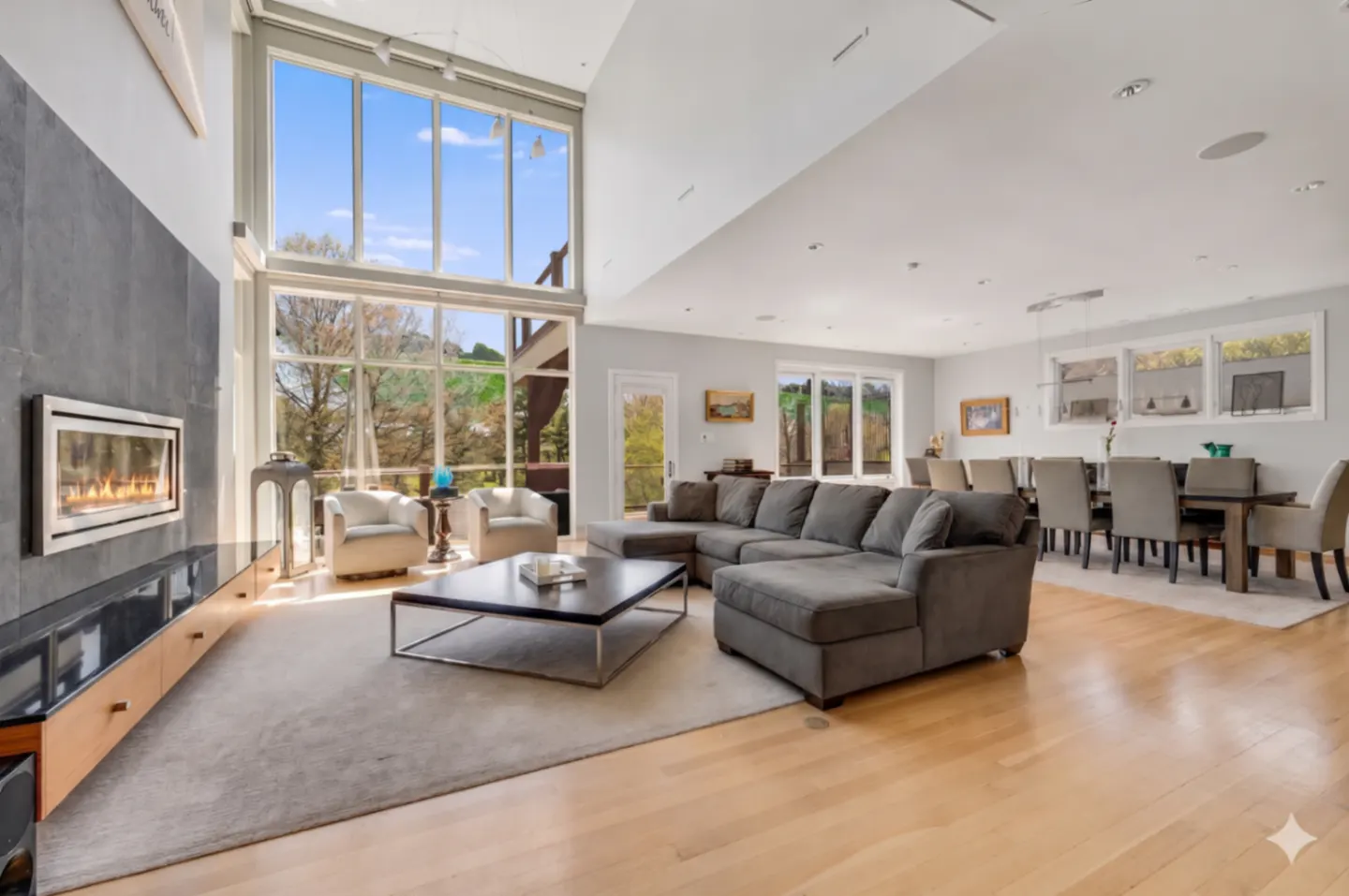 Open-concept living room with hardwood floors, a gray sectional sofa, and a modern fireplace. Large windows offer a view of the outdoors.