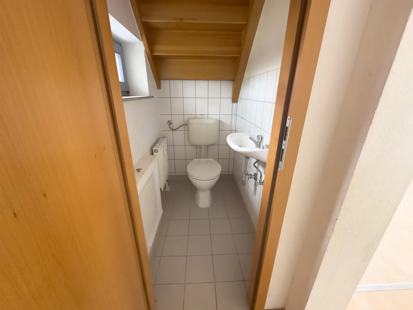 Small bathroom with white toilet, wall-mounted sink, and white tiled walls under a wooden staircase.