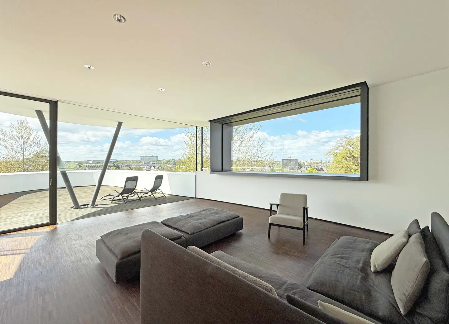 Bright, modern living room with a gray sofa, chair, and ottoman. Large windows offer city views and access to a balcony with chairs.