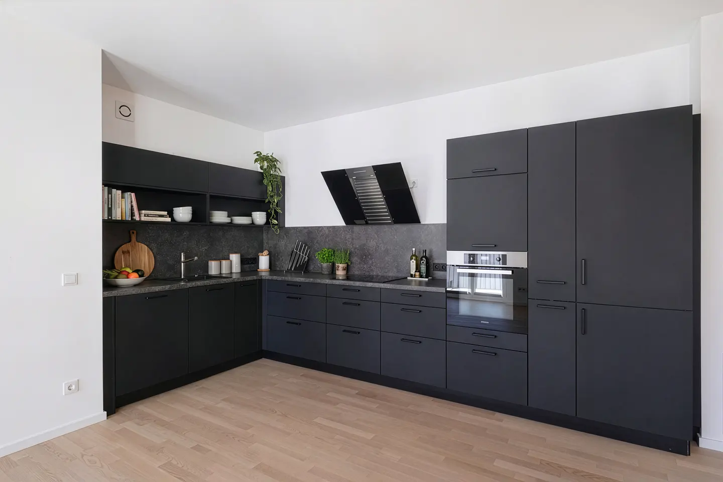 Modern kitchen with sleek, dark gray cabinets, stainless steel oven, and light wood floors. A black range hood is above the stove.